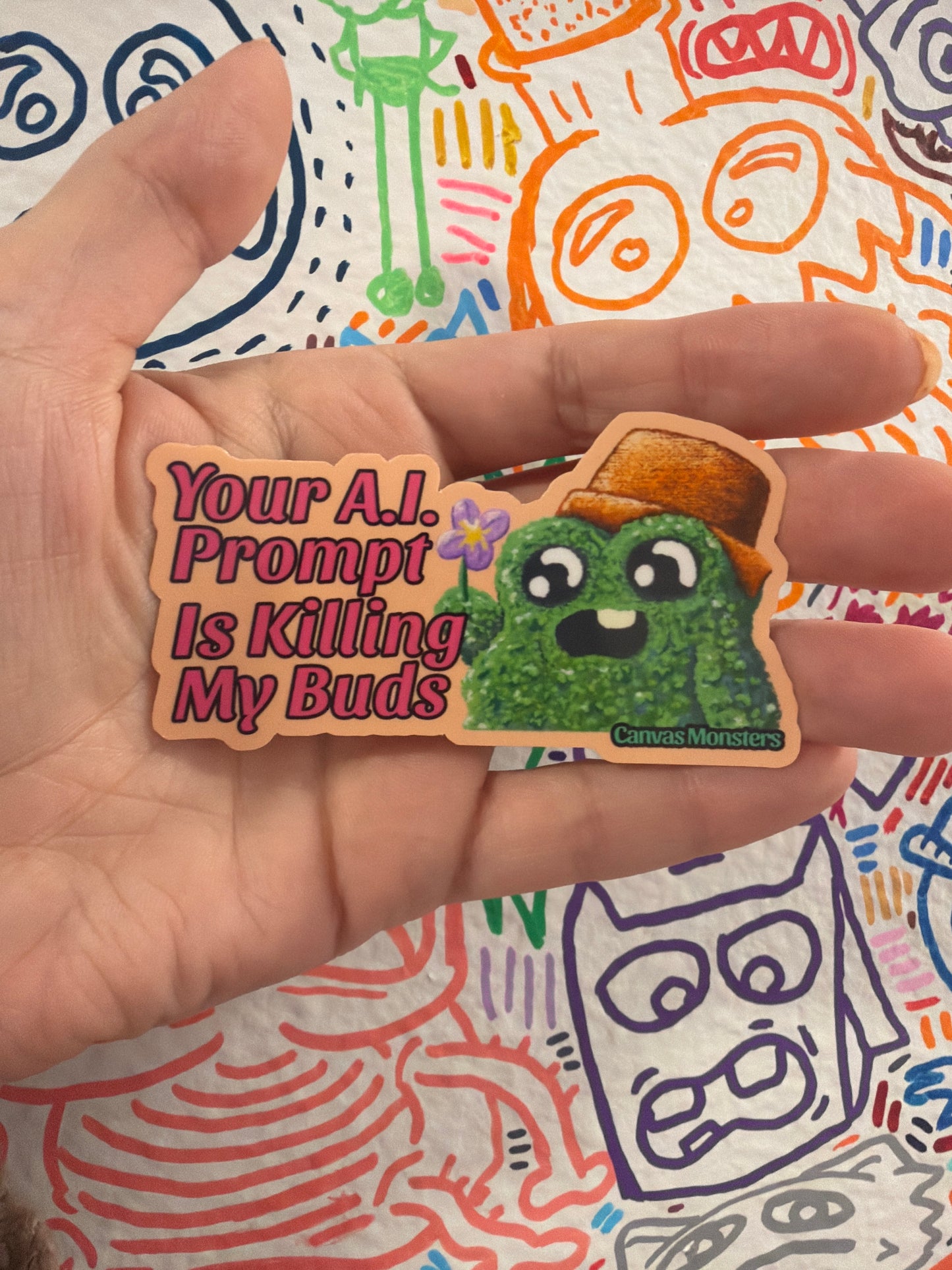 A hand holds a die-cut sticker featuring a green shrub cartoon character wearing an orange hat next to the pink text 'Your A.I. Prompt Is Killing My Buds"