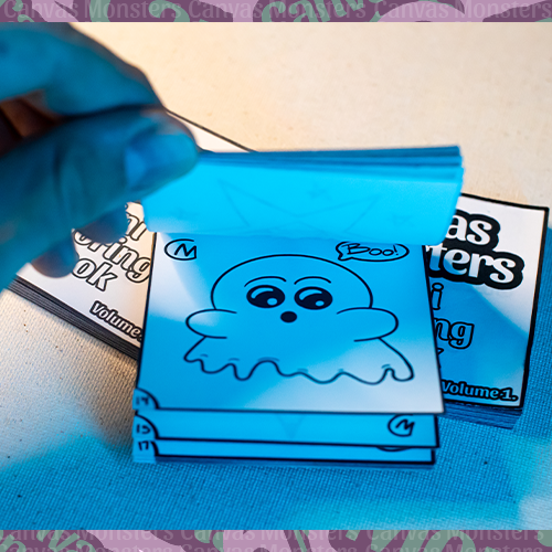 Hand flipping through the Canvas Monsters Mini Coloring Book Volume 1 on a white surface. Photo shown is a cartoon ghost.