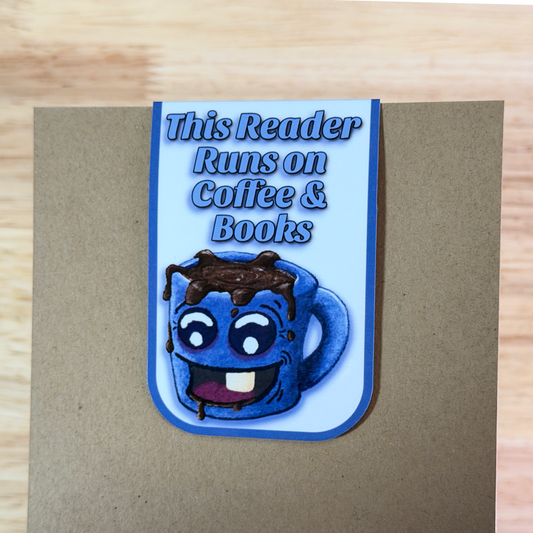 Magnetic bookmark with a blue cartoon coffee cup and text 'This Reader Runs on Coffee & Books' on a wooden surface.