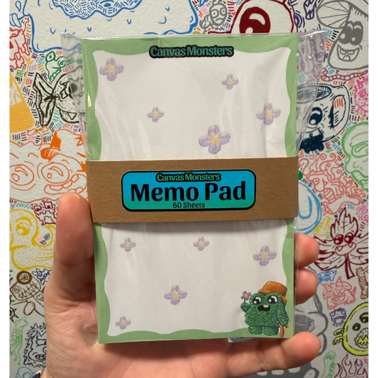 A hand holding a package of 4"x6" Canvas Monsters Memo Pad 60 sheets, featuring a white paper pad with light purple flower patterns and a brown paper band label, set against a colorful, doodle-filled background.