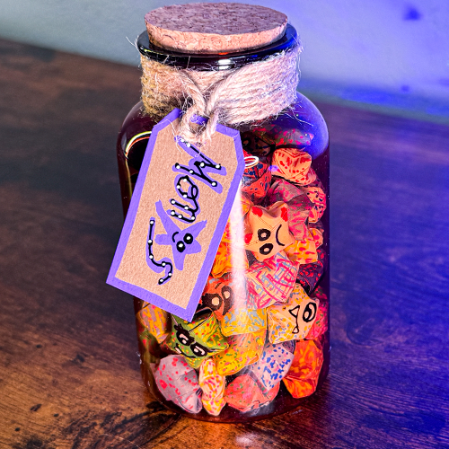 A glass jar filled with various colorful paper stars, sealed with a cork stopper and adorned with a decorative tag that reads 'Monstars - A Jar of Monsters'.