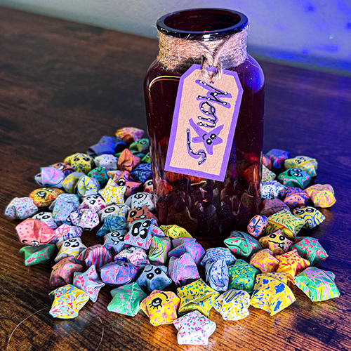 A glass jar surrounded by dozens of colorful paper stars, adorned with a decorative tag that reads 'Monstars - A Jar of Monsters'.