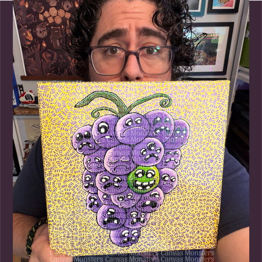 Mike, owner of Canvas Monsters, holding up a square canvas painting featuring a cluster of purple cartoon grapes with sad faces on a yellow patterned background. Mike is wearing glasses and a dark blue shirt, with framed artwork and shelves visible in the background.