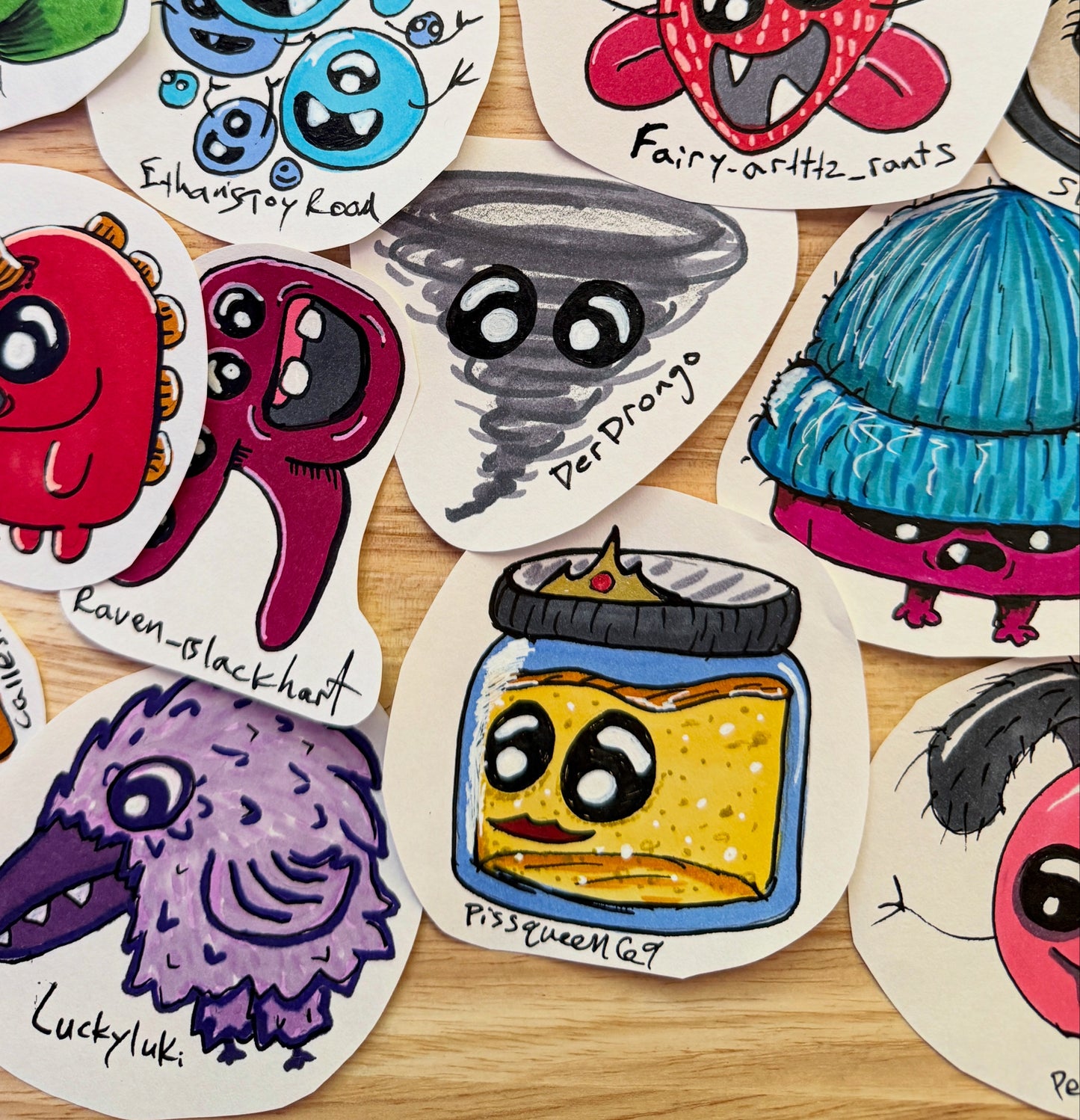 A collection of hand-drawn cartoon monster art on white paper, scattered on a light wood surface. The illustrations include colorful creatures like a red monster, a blue monster, a gray tornado, a purple raven, a yellow jar character, and others with handwritten labels.