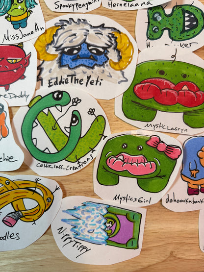 A collection of colorful, hand-drawn monster characters scattered on a light wood surface. Each drawing features a unique cartoon monster with a handwritten name underneath, such as 'Mystic Lasryn', and 'Mystics Girl'.