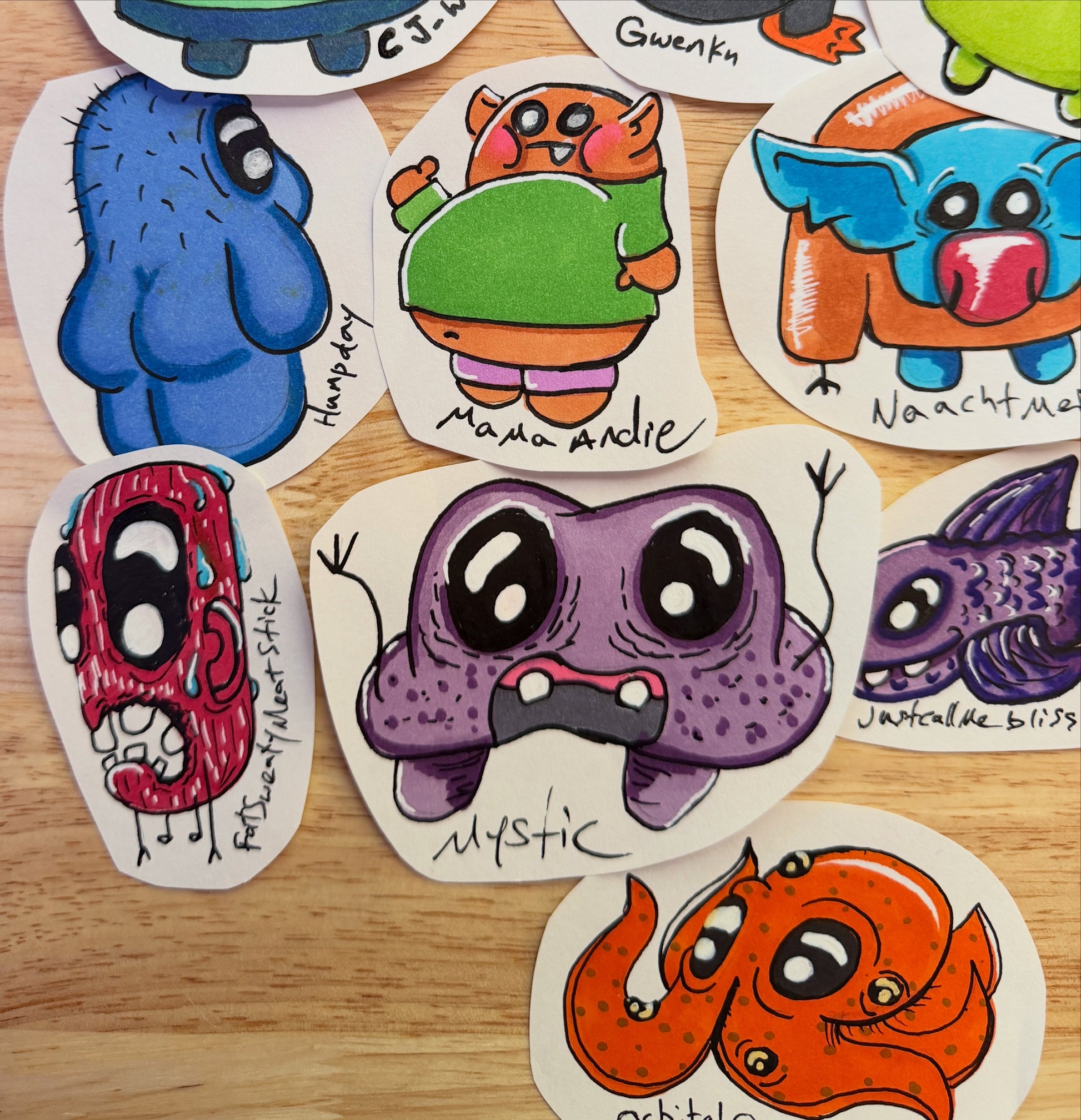 A collection of hand-drawn cartoon monsters on white paper, arranged on a light wood surface. The monsters feature various colorful, whimsical creatures with large eyes and unique designs, including blue, purple, orange, red, and green characters.