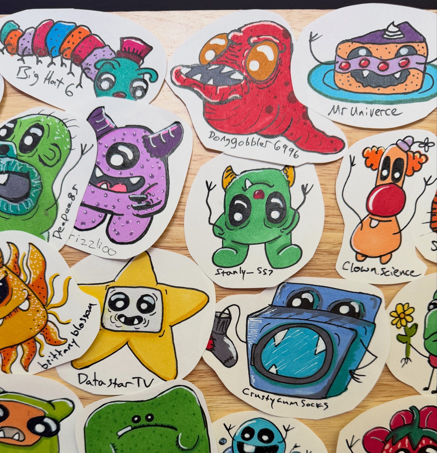 A collection of hand-drawn, die-cut sticker characters arranged on a light wood surface. The stickers feature whimsical, colorful cartoon designs including a green monster, a purple monster, a yellow star, a blue washing machine, and a green creature, each with handwritten labels.