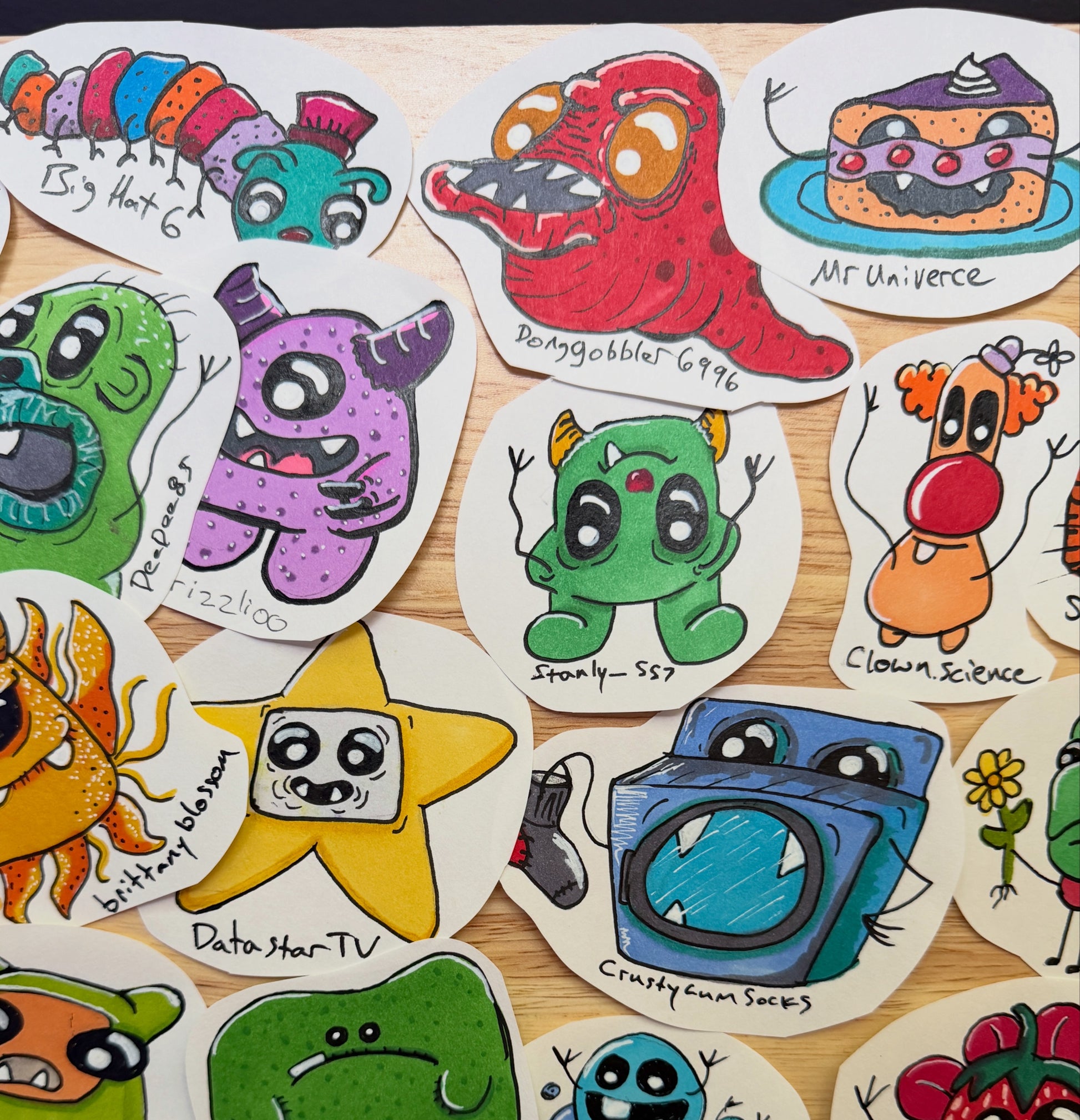 A collection of hand-drawn, die-cut sticker characters arranged on a light wood surface. The stickers feature whimsical, colorful cartoon designs including a green monster, a purple monster, a yellow star, a blue washing machine, and a green creature, each with handwritten labels.