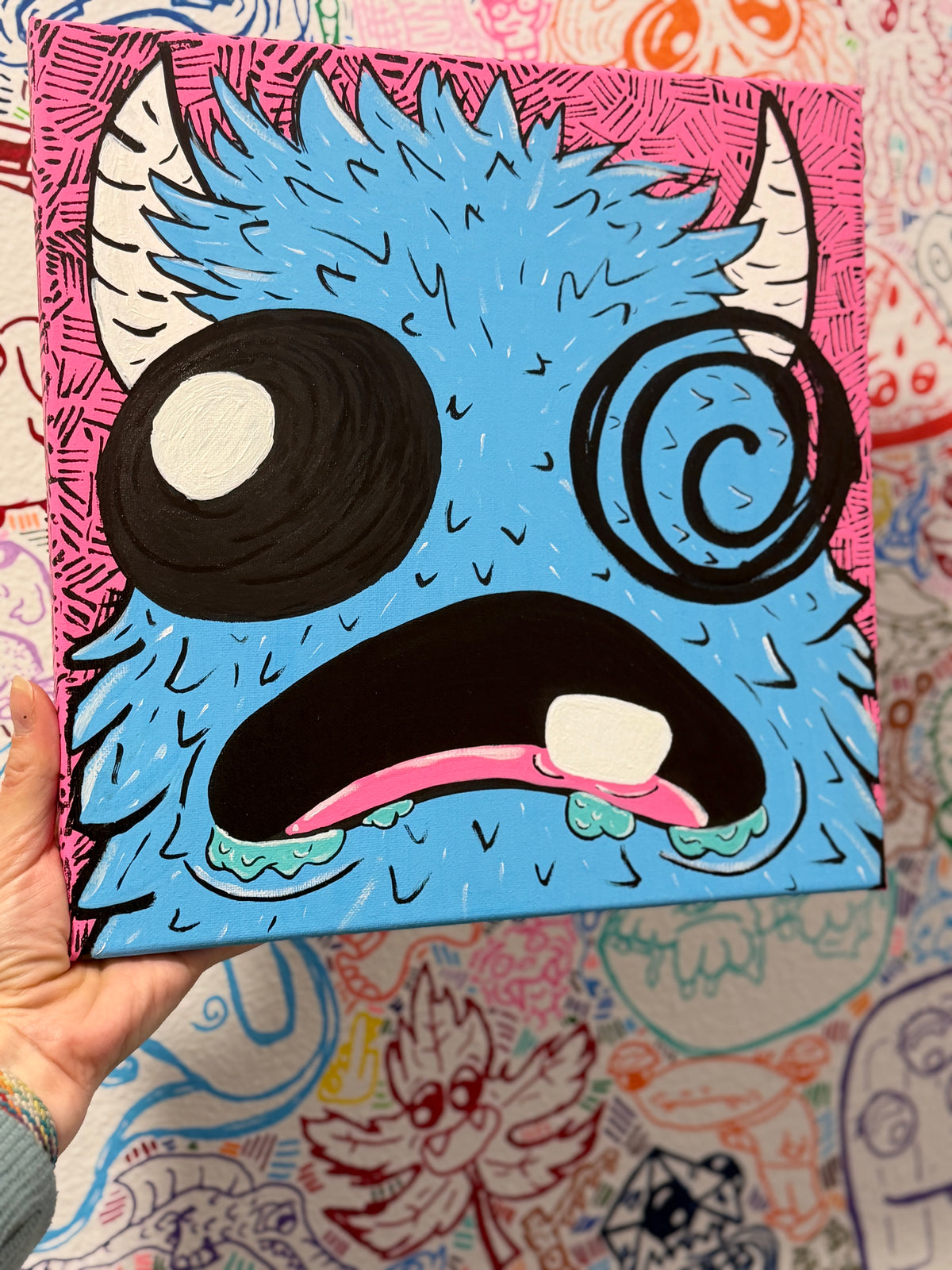Hand holding a painting of a blue monster character on a pink background, held against a patterned wall.