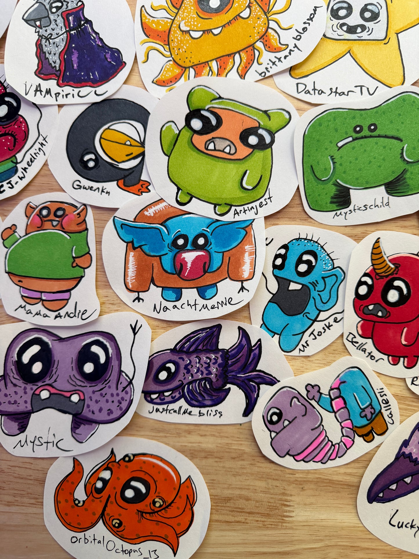 A collection of hand-drawn monster characters arranged on a light wood surface. Each unique cartoon creature features colorful body colors, large expressive eyes, and handwritten names underneath, such as 'Vampyre', 'Gruntle', 'Angst', 'Morphechild', 'Mama Andie', 'Naachtmerrie'.