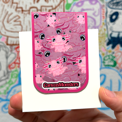 A hand holds a pink monster-themed sticker or card featuring multiple cartoon monster Axolotls and the text 'CanvasMonsters' at the bottom, against a colorful blurred background.