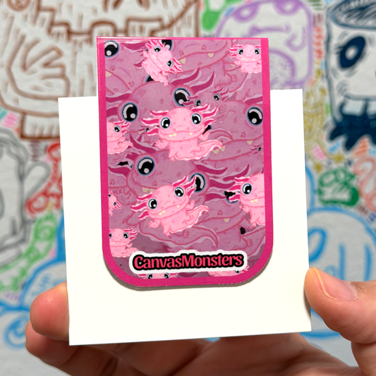 A hand holds a pink monster-themed sticker or card featuring multiple cartoon monster Axolotls and the text 'CanvasMonsters' at the bottom, against a colorful blurred background.