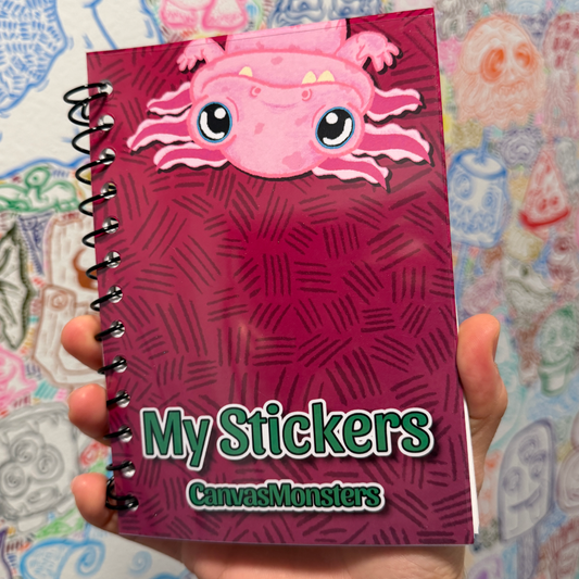 A hand holding a spiral-bound sticker book titled 'My Stickers' by CanvasMonsters. The cover is dark magenta with a cartoon pink axolotl at the top and green title text.