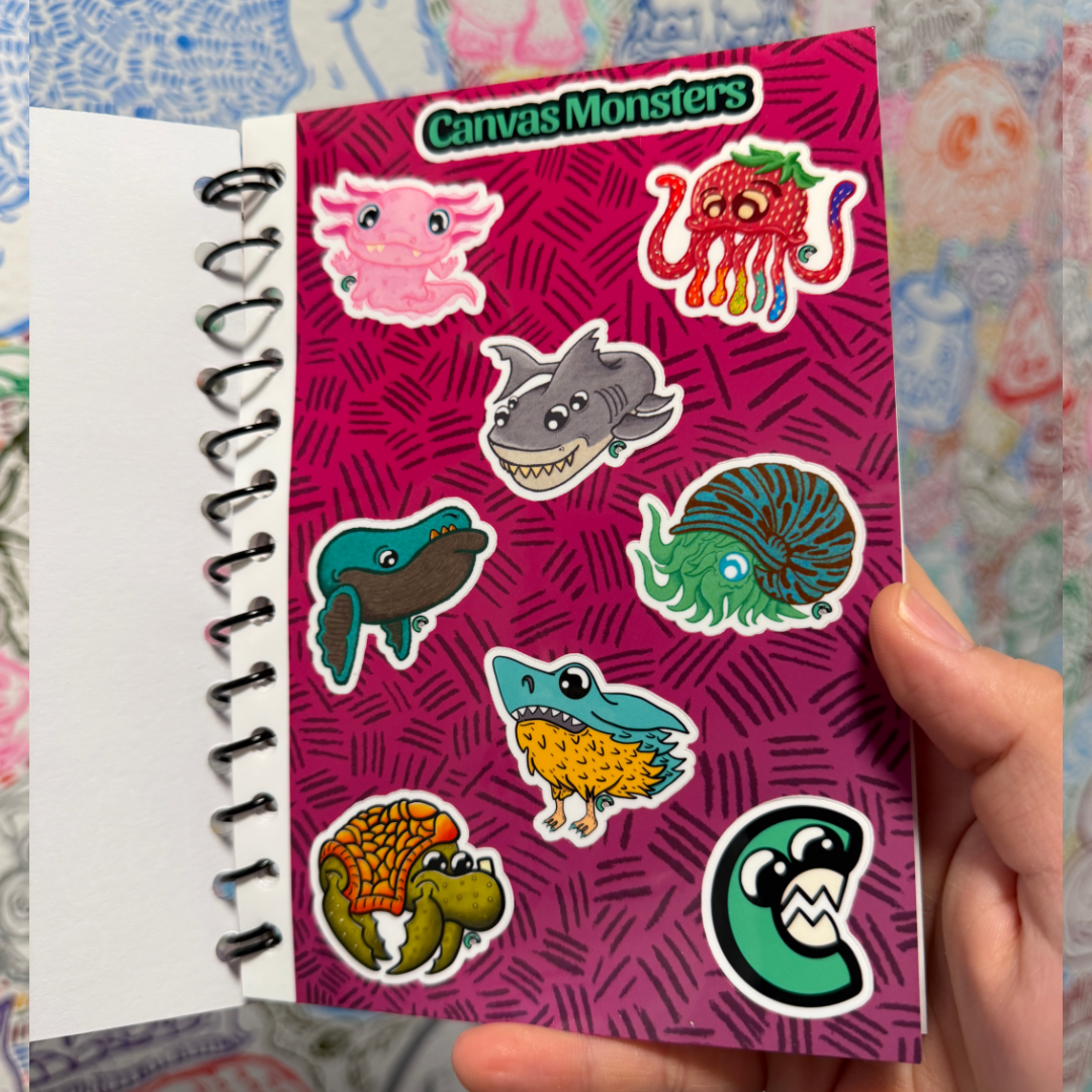 A hand holds a small spiral-bound notebook showing a sticker sheet with cartoon monster illustrations. The sheet has a bright pink background with the title 'Canvas Monsters' at the top, and several colorful monster stickers are visible.