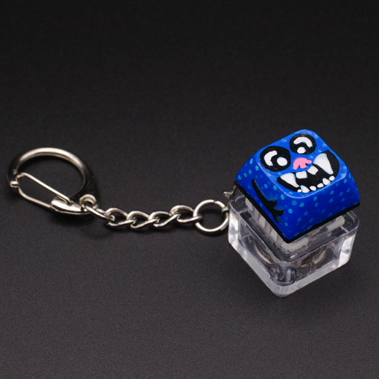 Photo of a key cap keychain on a black background. Cap is painted with a blue monster character.