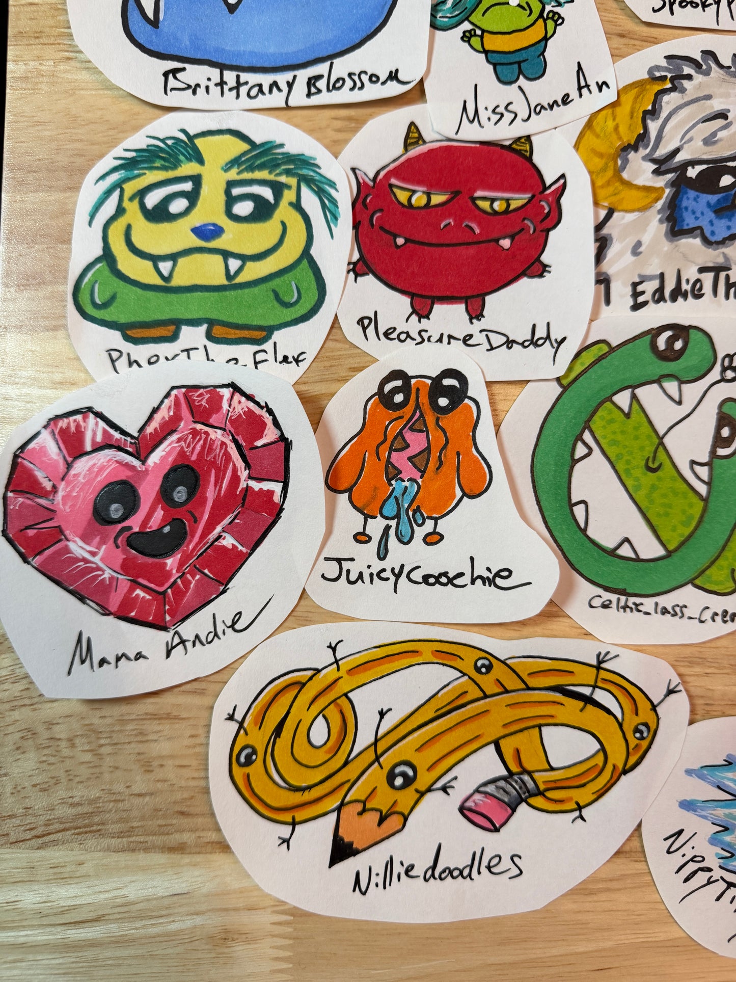 A collection of hand-drawn monster and character stickers arranged on a light wood surface. The stickers feature colorful cartoon designs including a green monster, a red demon, a pink heart, an orange dog, a yellow coiled snake, and others, each labeled with a playful name.