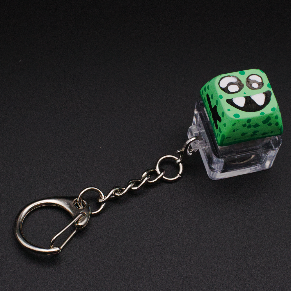 Photo of a key cap keychain on a black background. Cap is painted with a green monster character.