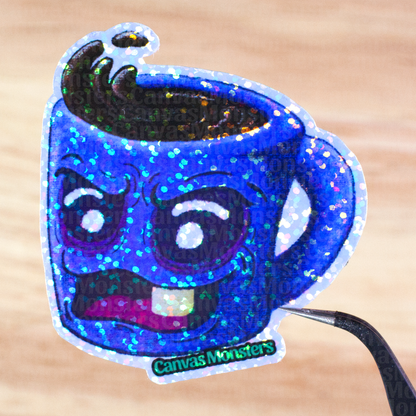 Glittery sticker of a blue mug with an angry face held above a wooden surface, branded 'Canvas Monsters'.