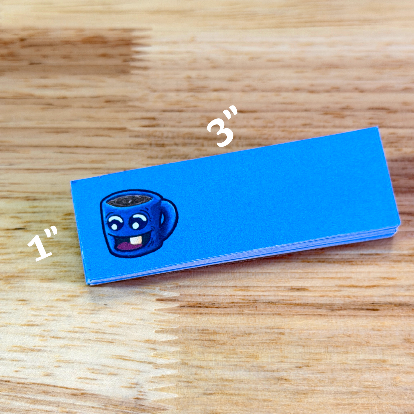1x3 sticky notepad with blue paper and cartoon cup of coffee on the left, sitting on a wooden surface.