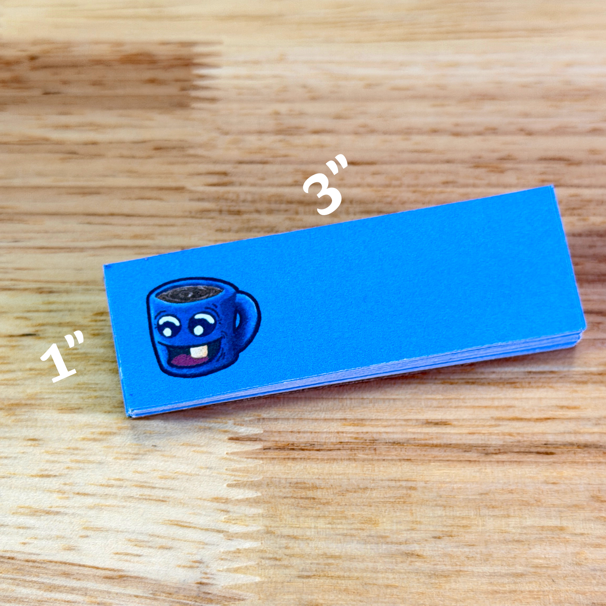 1x3 sticky notepad with blue paper and cartoon cup of coffee on the left, sitting on a wooden surface.