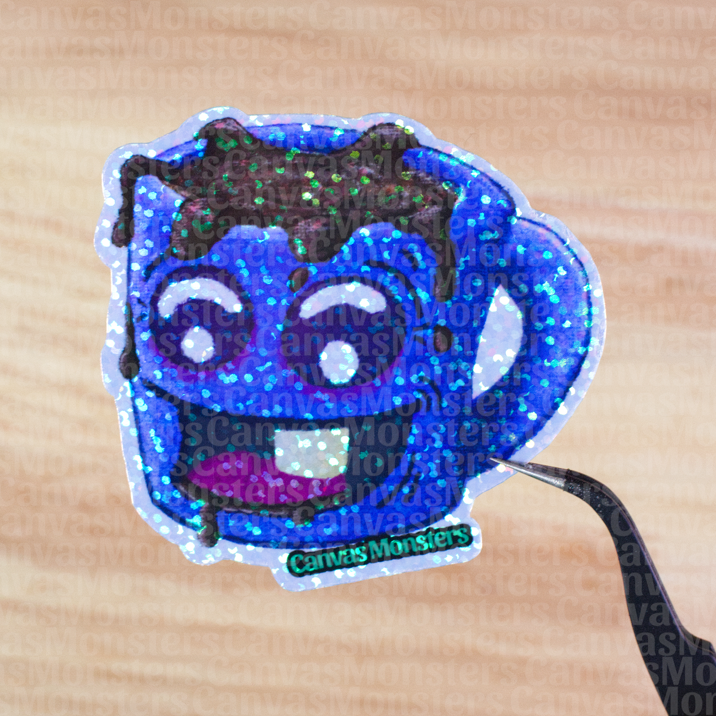 Glittery sticker of a blue mug with a happy face an coffee spilling out held above a wooden surface, branded 'Canvas Monsters'.