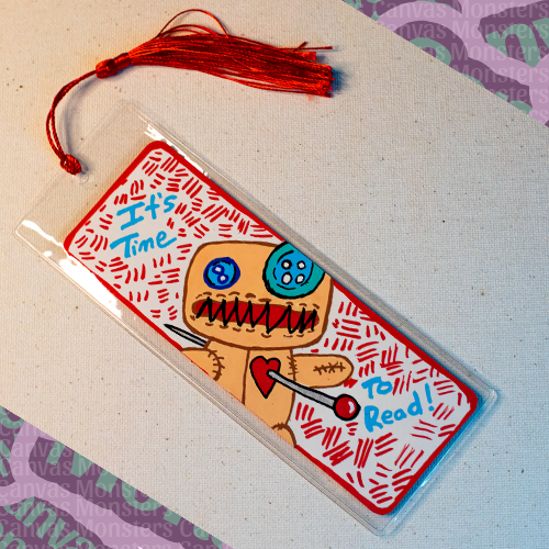 White paper bookmark with Canvas Monsters character "Dawl" painted on it with a red hash mark background and matching border, in a plastic sleeve and matching blue tassel. Text on the bookmark says 'It's Time To Read!'