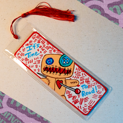White paper bookmark with Canvas Monsters character "Dawl" painted on it with a red hash mark background and matching border, in a plastic sleeve and matching blue tassel. Text on the bookmark says 'It's Time To Read!'