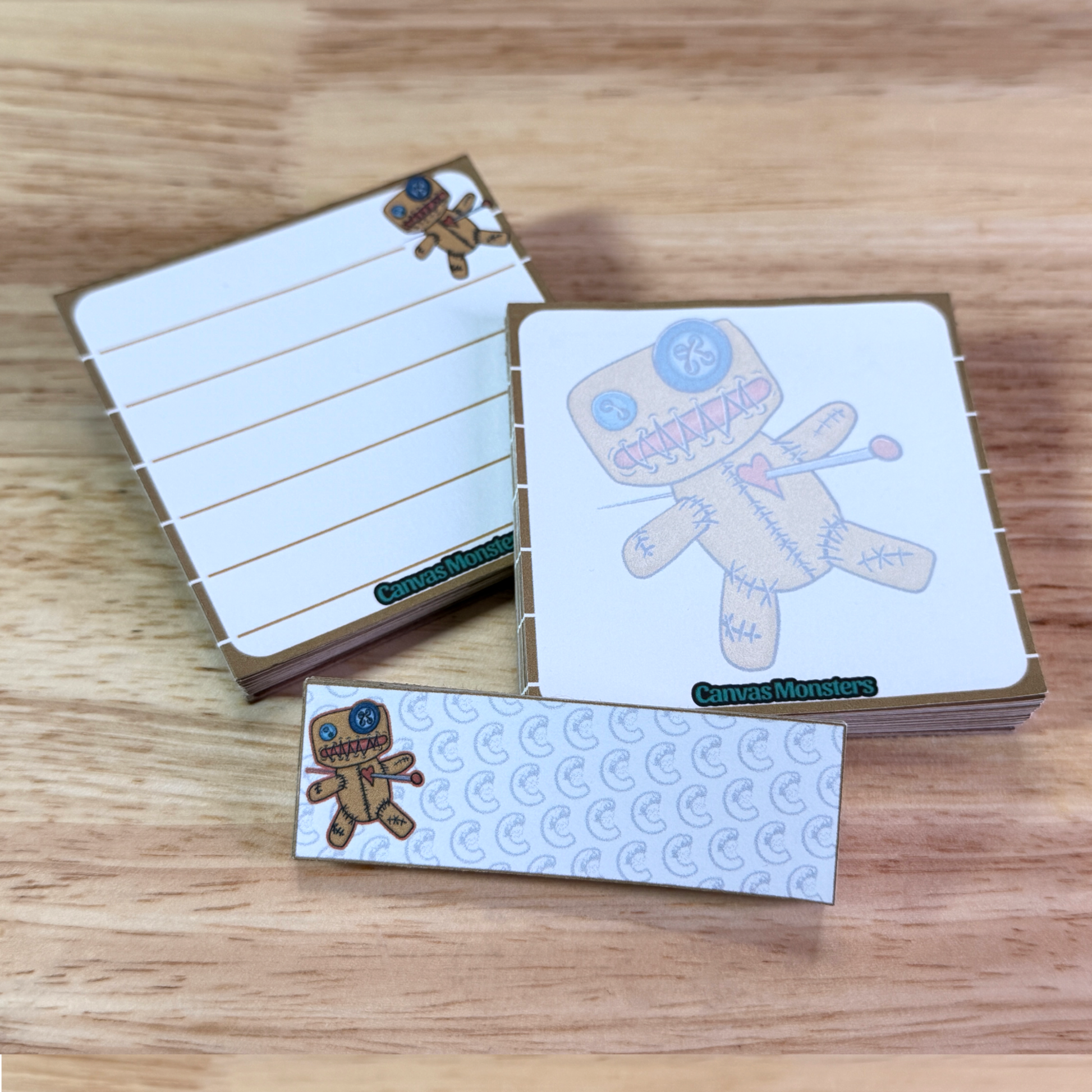 Full set of stationary, including a 3x3 memo pad, a 3x3 sticky notepad, and a 1x3 sticky notepad. Each with an illustration of a cartoon voodoo doll, all sitting on a wooden surface.