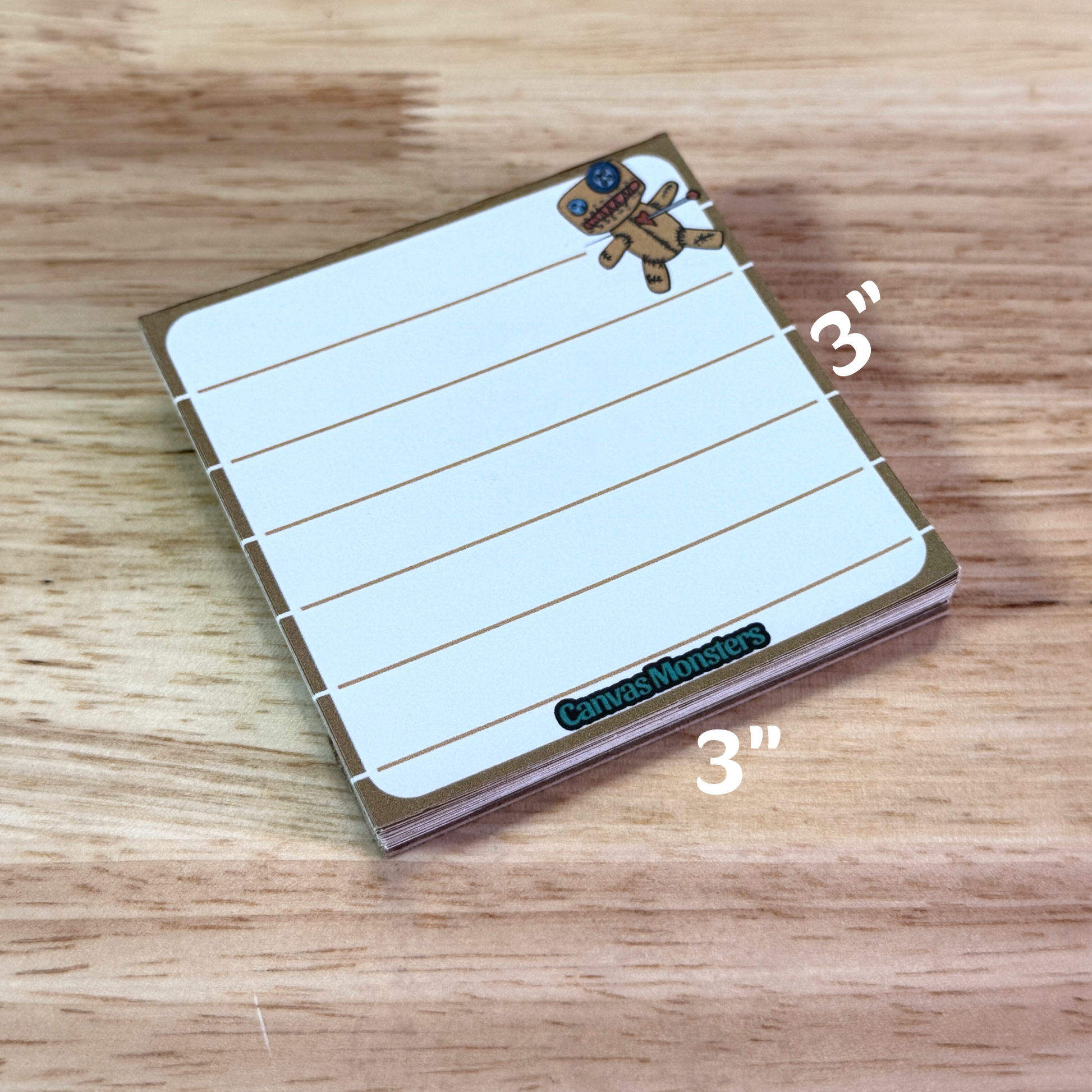 3x3 inch lined memo pad with a cartoon voodoo character in the top right corner and 'Canvas Monsters' branding at the bottom, sitting on a wooden surface.