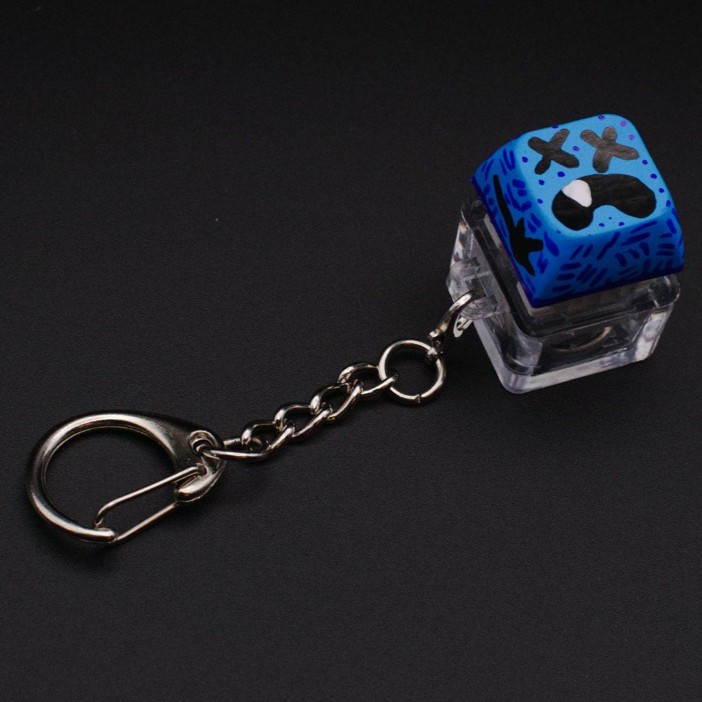 Photo of a key cap keychain on a black background. Cap is painted with a blue monster character.