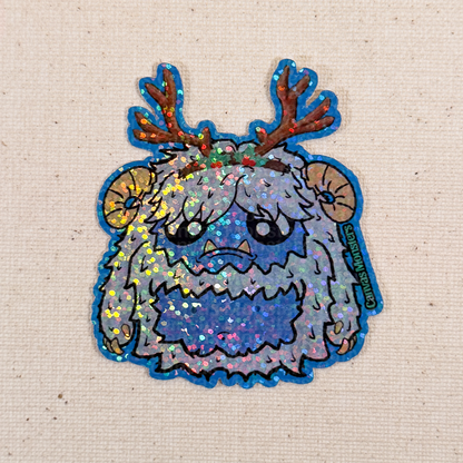 Glitter sticker of a cartoon yeti creature with white fur, blue belly, and antlers on a textured beige background.