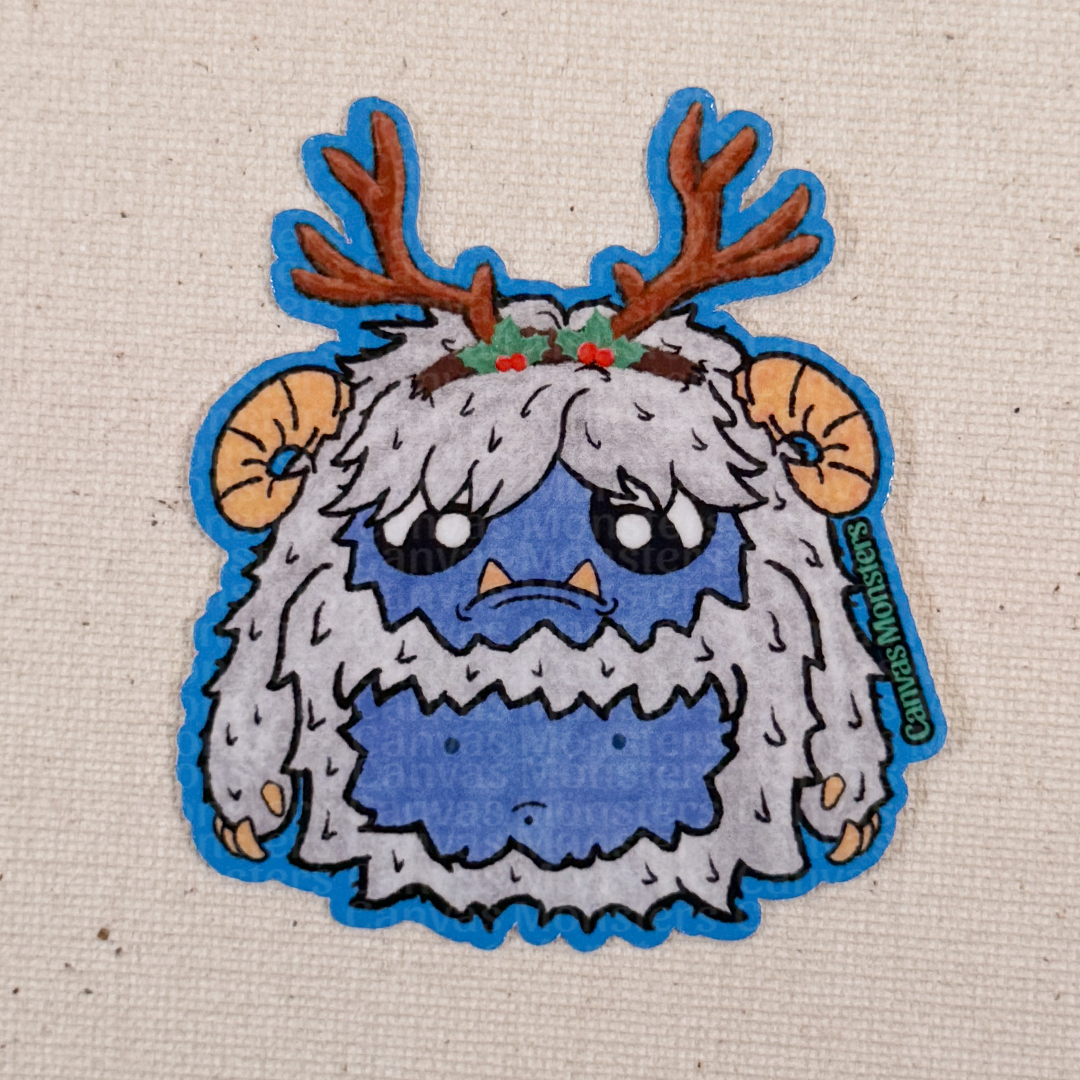 Glossy sticker of a cartoon yeti creature with white fur, blue belly, and antlers on a textured beige background.