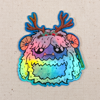 Holofoil sticker of a cartoon yeti creature with white fur, blue belly, and antlers on a textured beige background.