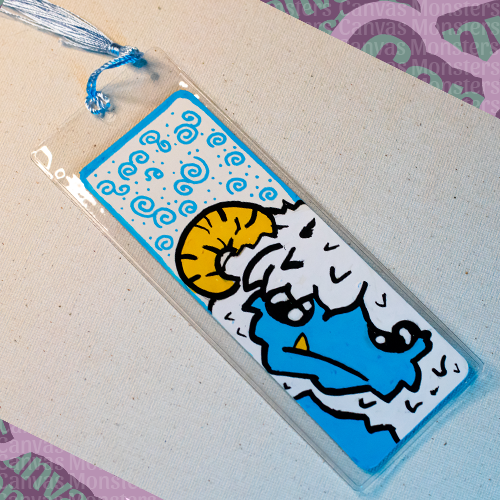 White paper bookmark with Canvas Monsters character "Eddie the Yeti" painted on it with a blue swirl background and border, in a plastic sleeve and matching blue tassel.