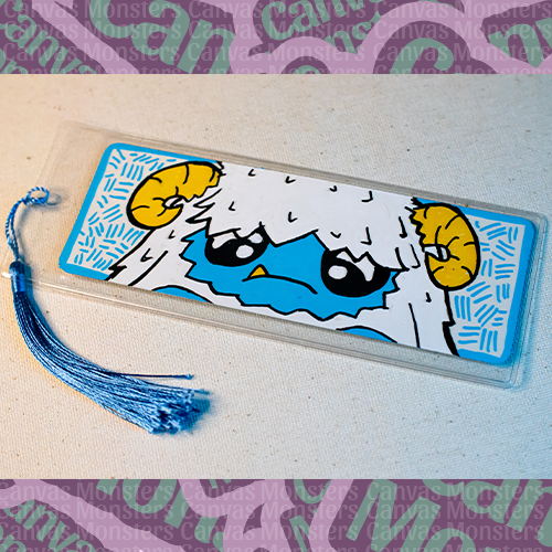 White paper bookmark with Canvas Monsters character "Eddie the Yeti" painted on it with a blue hash mark background and border, in a plastic sleeve and matching blue tassel.