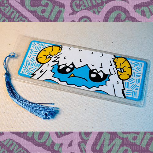 White paper bookmark with Canvas Monsters character "Eddie the Yeti" painted on it with a blue hash mark background and border, in a plastic sleeve and matching blue tassel.