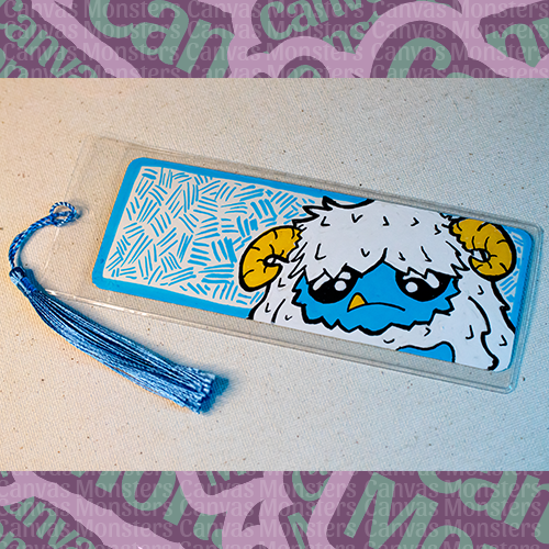 White paper bookmark with Canvas Monsters character "Eddie the Yeti" painted on it with a blue hash mark background and border, in a plastic sleeve and matching blue tassel.