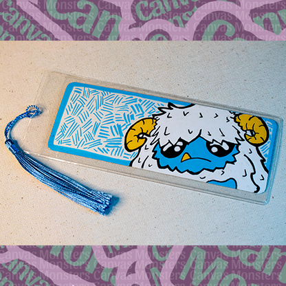 White paper bookmark with Canvas Monsters character "Eddie the Yeti" painted on it with a blue hash mark background and border, in a plastic sleeve and matching blue tassel.