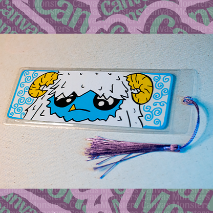White paper bookmark with Canvas Monsters character "Eddie the Yeti" painted on it with a blue swirl background and border, in a plastic sleeve and matching blue and purple tassel.