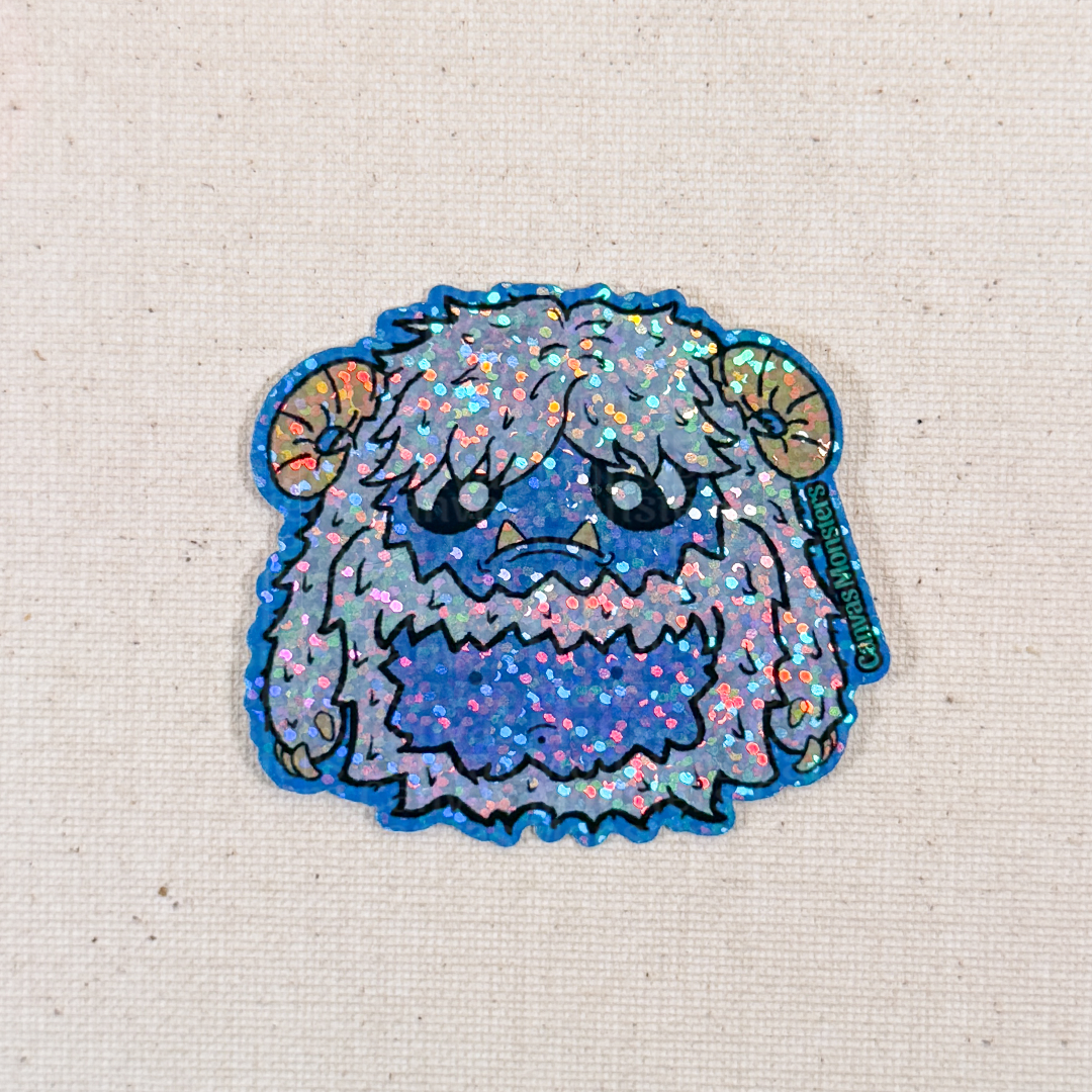 Glitter sticker of a cartoon yeti creature with white fur and horns on a textured beige background
