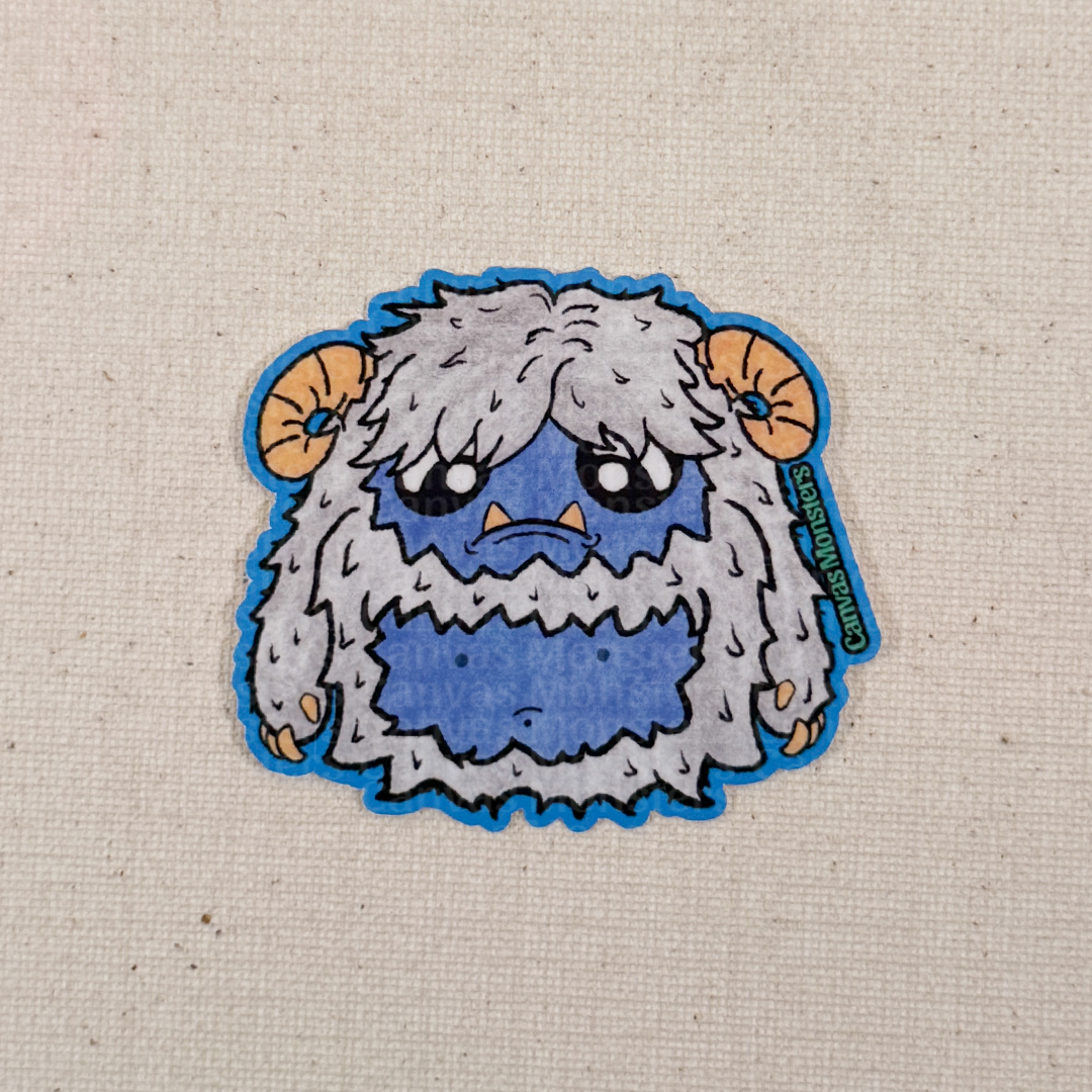 Glossy sticker of a cartoon yeti creature with white fur and horns on a textured beige background