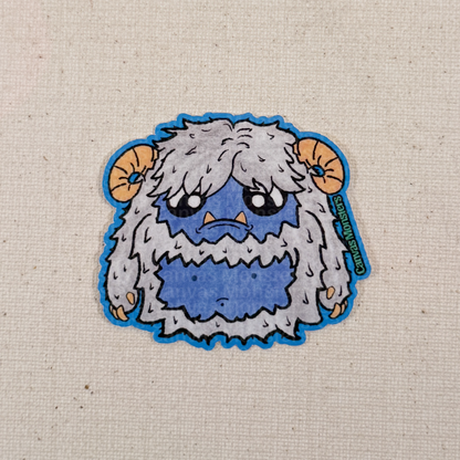 Glossy sticker of a cartoon yeti creature with white fur and horns on a textured beige background