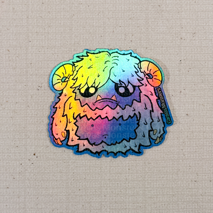 Holofoil sticker of a cartoon yeti creature with white fur and horns on a textured beige background