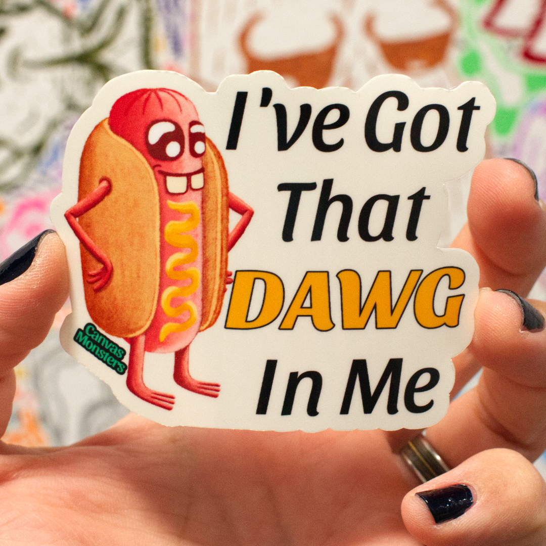 A hand holds a die-cut sticker featuring a cartoon hot dog with the text 'I've Got That DAWG In Me'. The sticker has a white background, black text, and a yellow/orange hot dog illustration with red details.