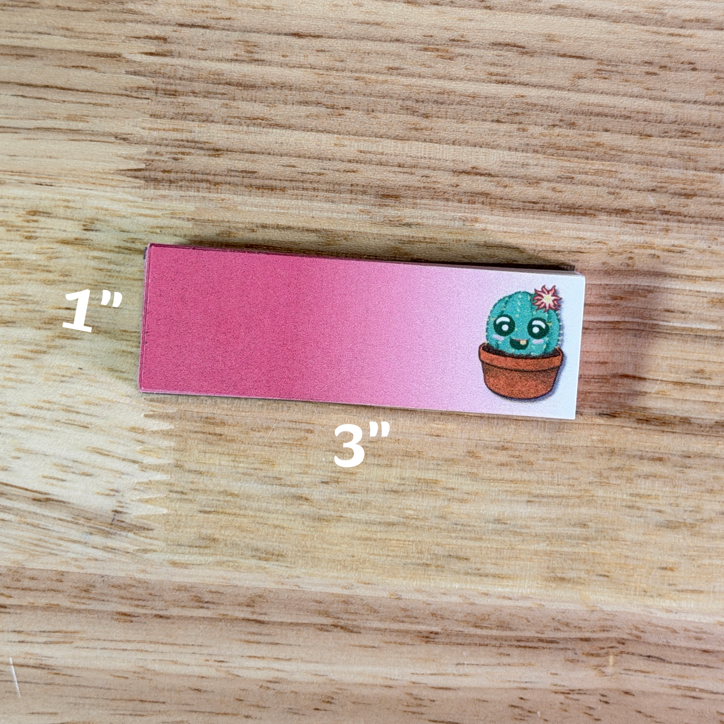 A 1x3 inch memo pad with pink to white gradient paper and a cartoon cactus illustrated on the right side, sitting on a wooden surface.