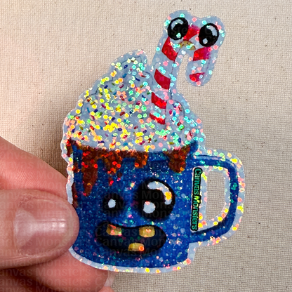 Glittery blue mug with whipped cream and candy cane held by a hand on a beige background
