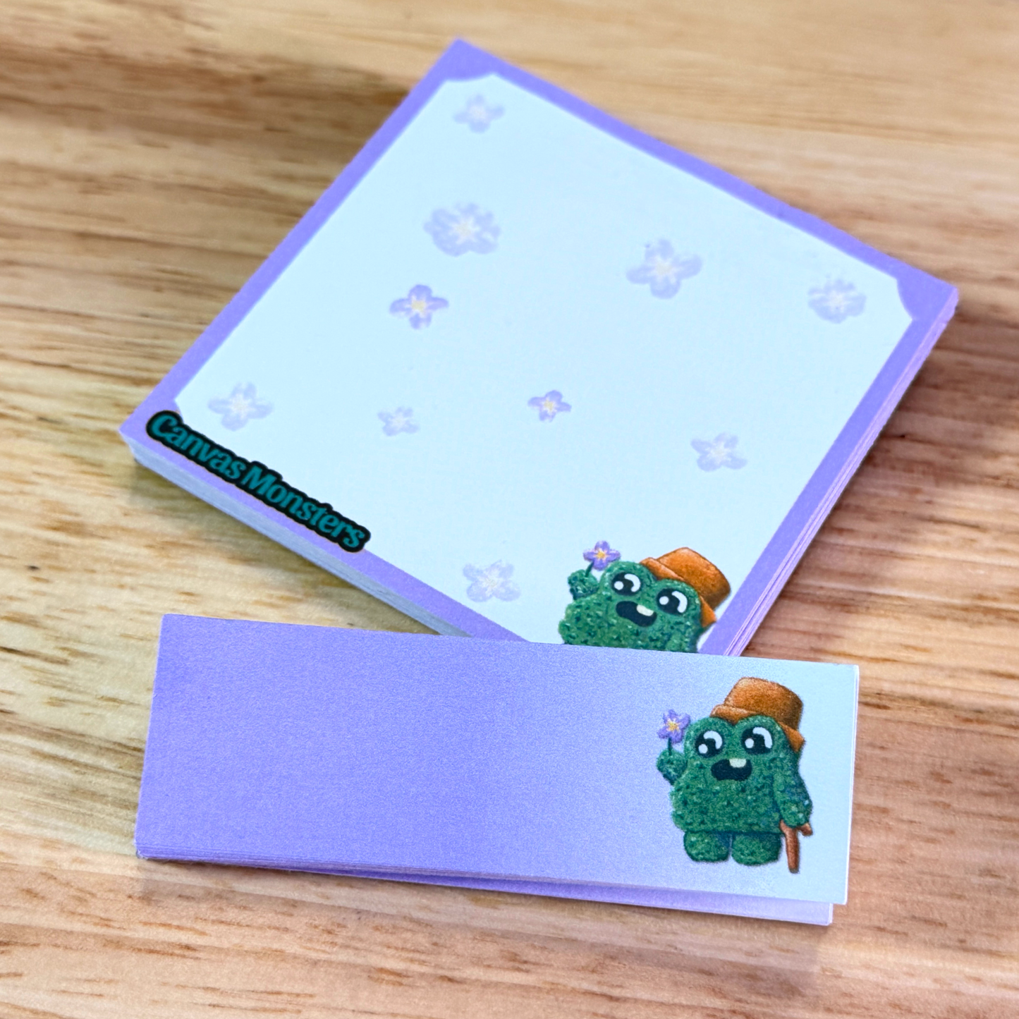 A matching set of sticky notepads with purple and white, and a cartoon shrub character, sitting on a wooden background.