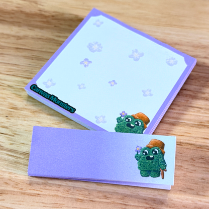 A matching set of sticky notepads with purple and white, and a cartoon shrub character, sitting on a wooden background.