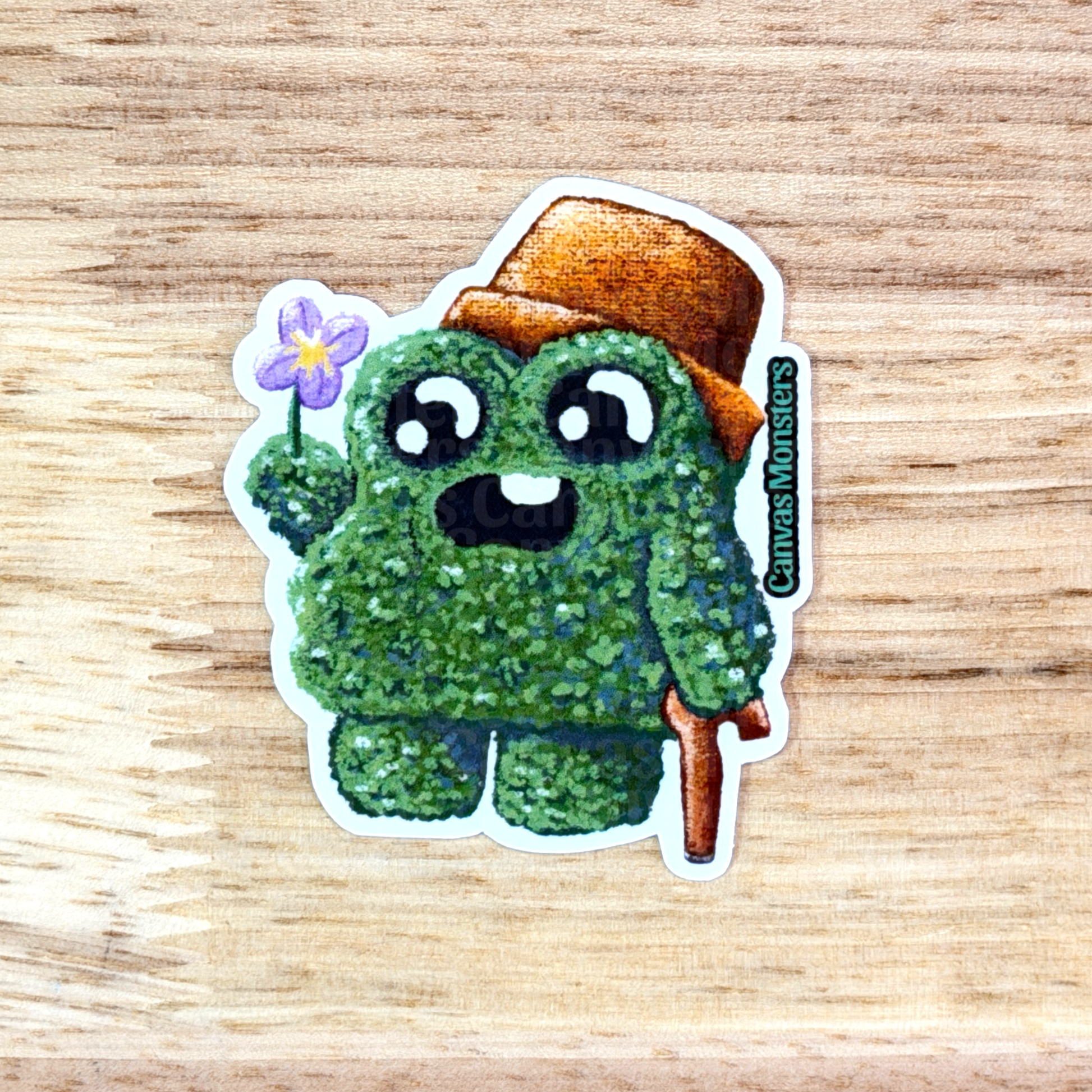 Sticker of a green creature with a brown hat holding a flower on a wooden surface