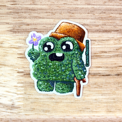 Sticker of a green creature with a brown hat holding a flower on a wooden surface